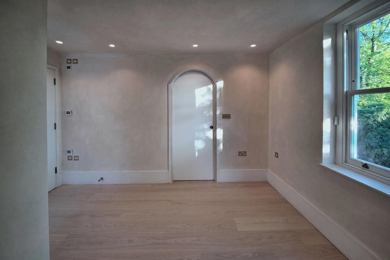 A limewashed room with a white arched door at the centre catching the sun, brass sockets, a pale plank floor and a sash window to trees on the right.